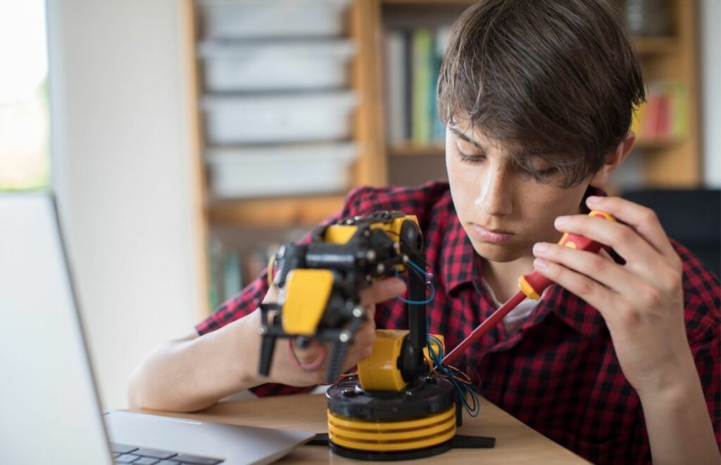 A young man building a robotic arm at his desk with some books in the background | best robotics books for beginners